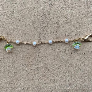 May include: A gold chain with white pearl beads and green flower charms. The chain has a clasp on each end.