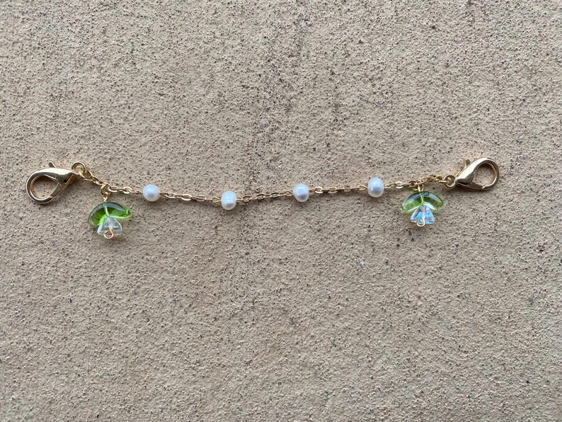 May include: A gold chain with white pearl beads and green flower charms. The chain has a clasp on each end.