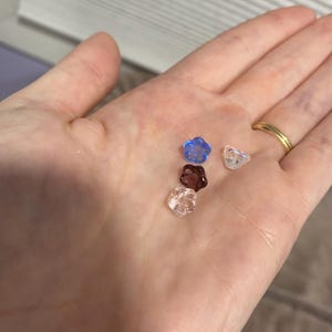 May include: Four small glass flower-shaped beads in different colours: light blue, clear, pink, and dark purple.