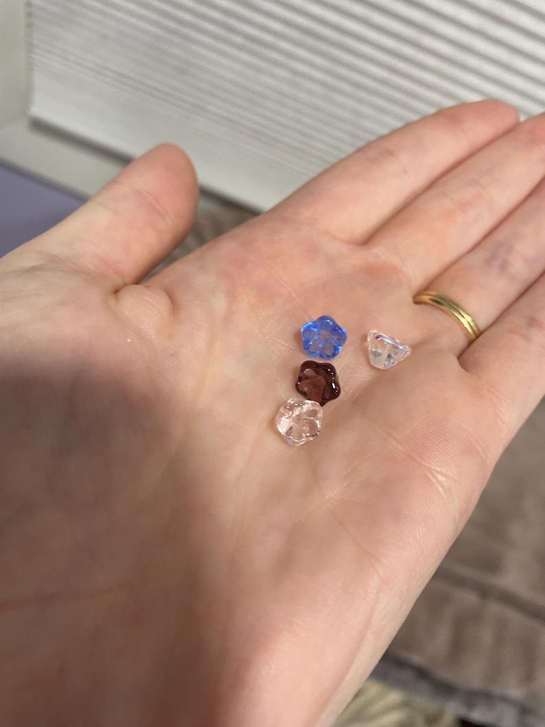 May include: Four small glass flower-shaped beads in different colours: light blue, clear, pink, and dark purple.