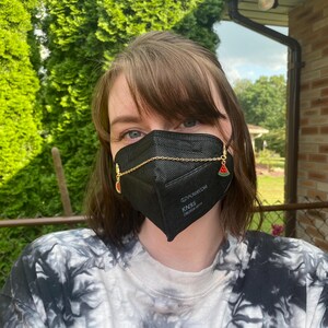 May include: A black KN95 face mask with a gold chain and watermelon charms. The mask has the text "EZPOWECOM KN95" printed on it. The person is wearing a tie-dye shirt with a pink heart graphic.