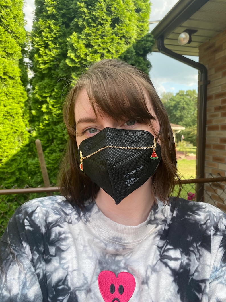 May include: A black KN95 face mask with a gold chain and watermelon charms. The mask has the text "EZPOWECOM KN95" printed on it. The person is wearing a tie-dye shirt with a pink heart graphic.