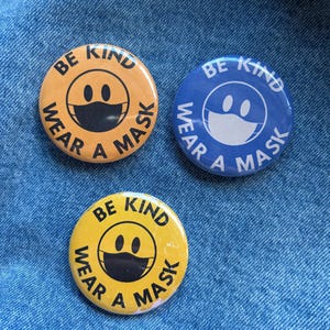 May include: Three round buttons with the text "BE KIND WEAR A MASK" around a smiley face wearing a mask. One button is yellow with black text and design, another is blue with white text and design, and the third is yellow with black text and design.