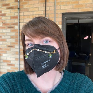 May include: A black face mask with a gold chain and floral charms attached. The mask has the text "KN95" and "GB2626-2019" printed on it.