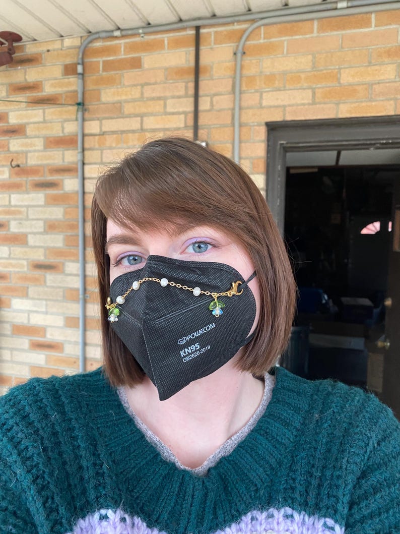 May include: A black face mask with a gold chain and floral charms attached. The mask has the text "KN95" and "GB2626-2019" printed on it.