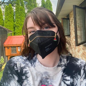 May include: A black KN95 face mask with a gold chain and watermelon charms. The mask has the text "POURKOM KN95" and "EN149:2001+A1:2009" printed on it. The person is wearing a tie-dye shirt.