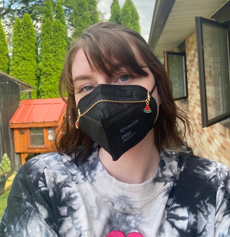 May include: A black KN95 face mask with a gold chain and watermelon charms. The mask has the text "POURKOM KN95" and "EN149:2001+A1:2009" printed on it. The person is wearing a tie-dye shirt.
