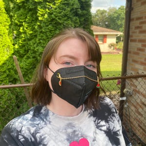 May include: A black face mask with gold chain and watermelon and strawberry charms. The mask is worn over the mouth and nose. The person is wearing a black and white tie-dye sweatshirt with a pink heart graphic.