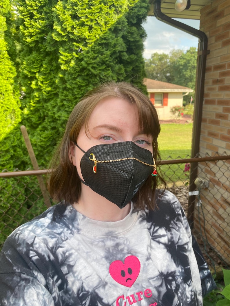 May include: A black face mask with gold chain and watermelon and strawberry charms. The mask is worn over the mouth and nose. The person is wearing a black and white tie-dye sweatshirt with a pink heart graphic.