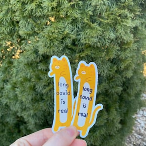 May include: Two yellow cat stickers with the text "long covid is real" on each. The stickers are held in a hand against a green leafy background.