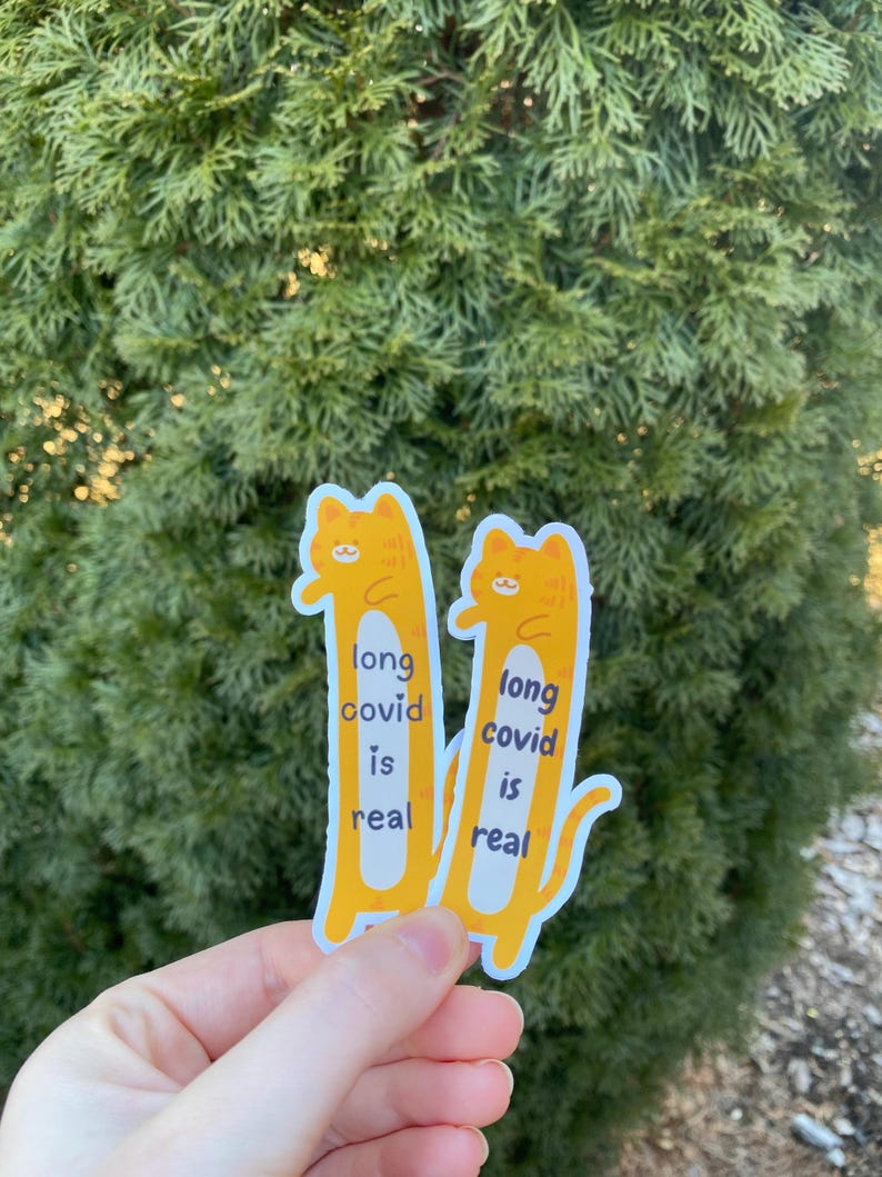 May include: Two yellow cat stickers with the text "long covid is real" on each. The stickers are held in a hand against a green leafy background.