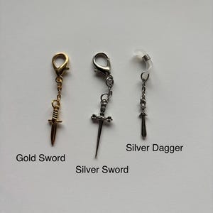 以下が含まれることがあります： 3つの剣をテーマにしたアクセサリー：金の剣チャーム、銀の剣チャーム、銀の短剣チャーム。各チャームは留め金またはメガネホルダーに取り付けられています。画像には「Gold Sword」、「Silver Sword」、「Silver Dagger」と書かれています。