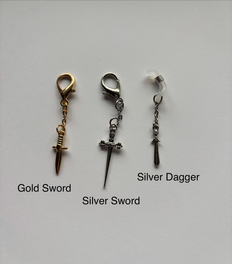 以下が含まれることがあります： 3つの剣をテーマにしたアクセサリー：金の剣チャーム、銀の剣チャーム、銀の短剣チャーム。各チャームは留め金またはメガネホルダーに取り付けられています。画像には「Gold Sword」、「Silver Sword」、「Silver Dagger」と書かれています。
