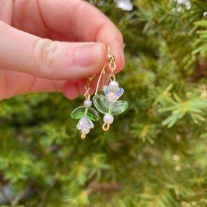 May include: A pair of gold dangle earrings featuring green and white glass beads shaped like flowers. The earrings are hanging from a person's ear.