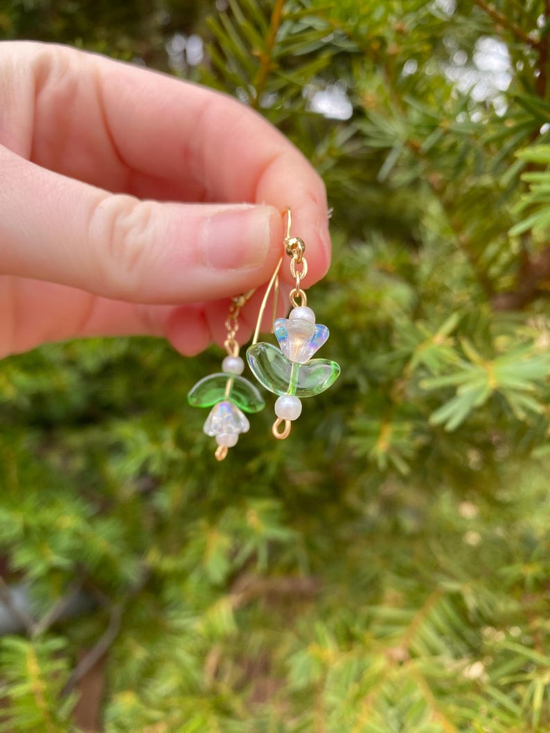 May include: A pair of gold dangle earrings featuring green and white glass beads shaped like flowers. The earrings are hanging from a person's ear.