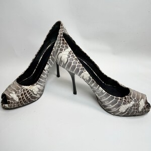 May include: A pair of peep-toe high heels with a snakeskin pattern in shades of gray and white. The heels are black and the inner soles are black. The shoes are displayed on a white background.