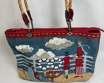May include: A blue tote bag with a red top and woven handles. The bag features an embroidered seaside scene with figures, clouds, and life preservers. The handles are made of woven rope and attached with red leather.