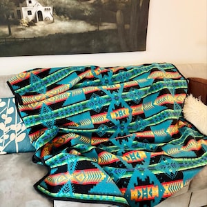 May include: A rectangular throw blanket featuring a vibrant geometric pattern in shades of turquoise, black, yellow, and red. The design consists of repeating diamond and arrow motifs arranged in horizontal bands across the fabric surface.