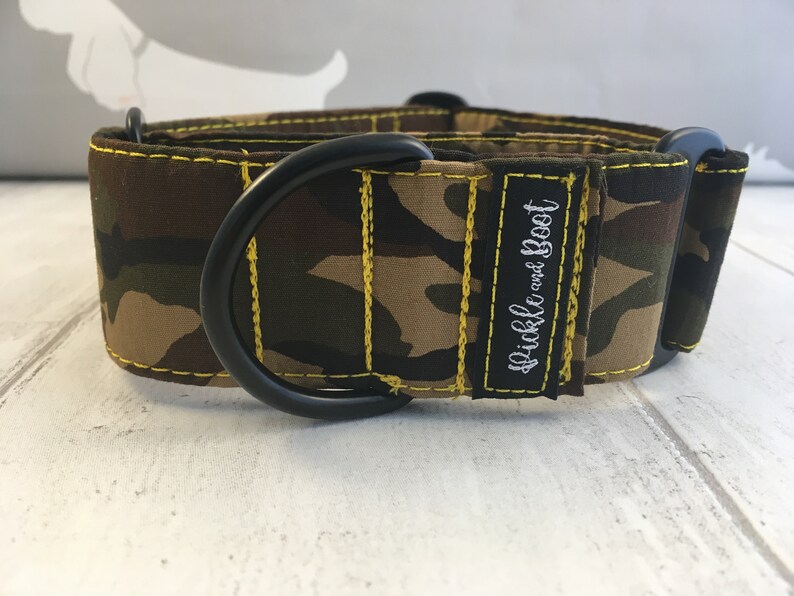 May include: A camouflage patterned dog collar with yellow stitching and a black metal D-ring. The collar has the text "Pickle and Boot" embroidered on it.