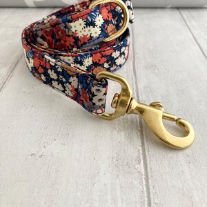 May include: A floral patterned dog leash with a brass clasp and hardware. The leash features a repeating pattern of red, white, and blue flowers on a navy blue background. The brass hardware includes a swivel clasp and a D-ring.
