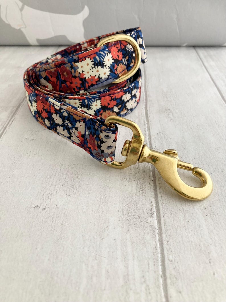 May include: A floral patterned dog leash with a brass clasp and hardware. The leash features a repeating pattern of red, white, and blue flowers on a navy blue background. The brass hardware includes a swivel clasp and a D-ring.
