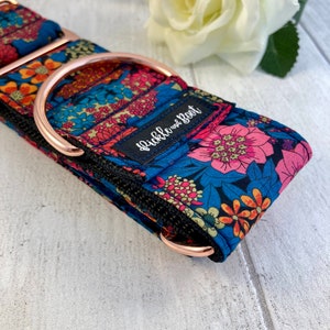 K&ouml;nnte beinhalten: Ein gemustertes Hundehalsband mit einem D-Ring und Beschl&auml;gen aus ros&eacute;goldfarbenem Metall. Das Halsband zeigt ein Blumenmuster in Rosa, Orange und Blau auf dunkelblauem Grund. Der Text "Pickle and Root" ist auf dem Halsband zu sehen.