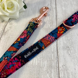 K&ouml;nnte beinhalten: Ein Hundeleinen- und Halsband-Set mit floralem Muster. Das Design zeigt rosa, blaue, orangefarbene und gr&uuml;ne Blumen auf einem dunkelblauen Hintergrund. Die Leine hat einen ros&eacute;goldenen Verschluss und der Markenname "Pickle and Boot" ist sichtbar.