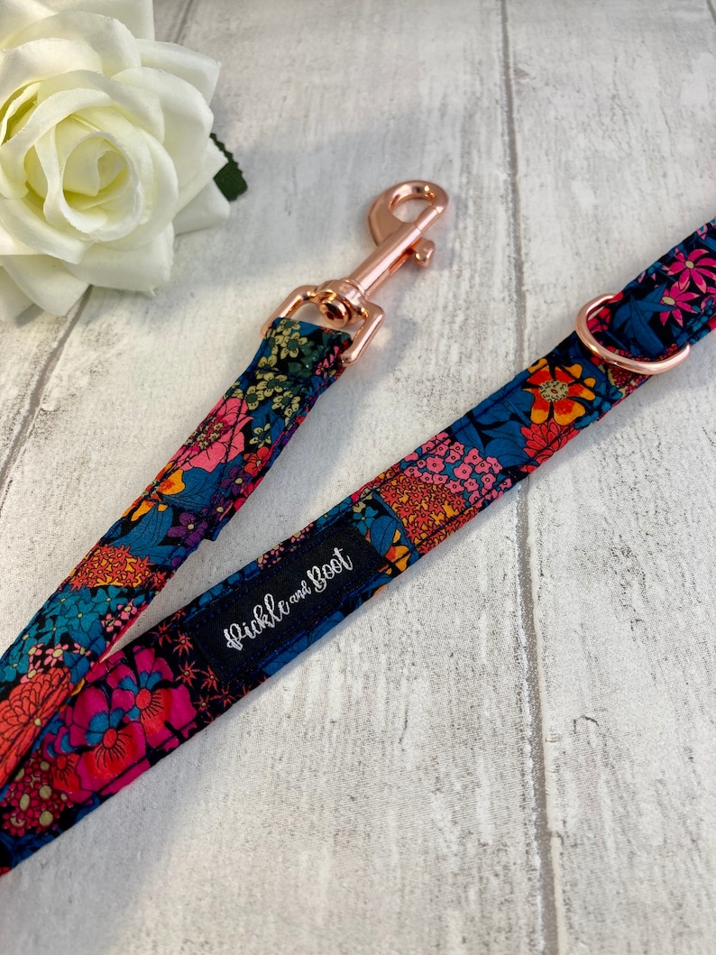 K&ouml;nnte beinhalten: Ein Hundeleinen- und Halsband-Set mit floralem Muster. Das Design zeigt rosa, blaue, orangefarbene und gr&uuml;ne Blumen auf einem dunkelblauen Hintergrund. Die Leine hat einen ros&eacute;goldenen Verschluss und der Markenname "Pickle and Boot" ist sichtbar.