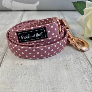 May include: A rolled dog lead in mauve with white polka dots. It features a rose gold clasp and a black label with the text "Pickle and Boot" in white script. The lead is designed for a pet.