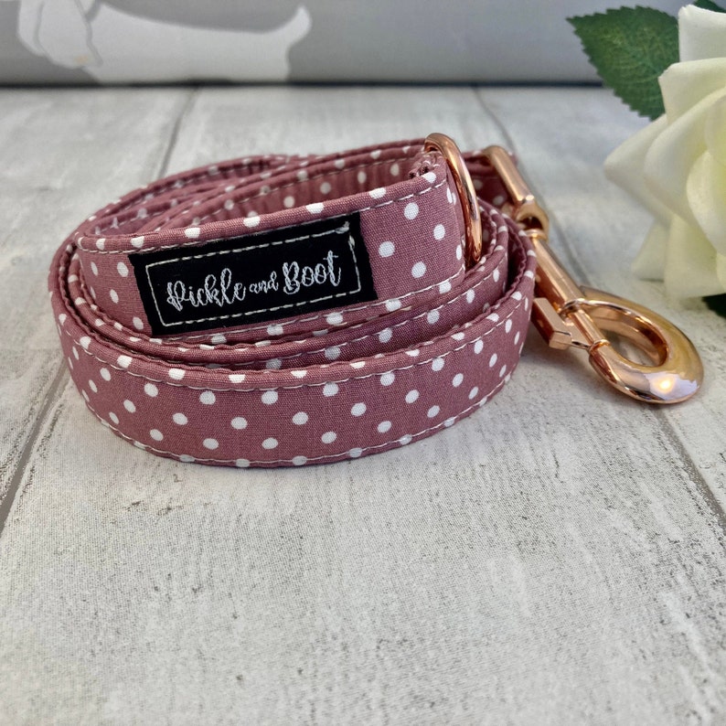 May include: A rolled dog lead in mauve with white polka dots. It features a rose gold clasp and a black label with the text "Pickle and Boot" in white script. The lead is designed for a pet.