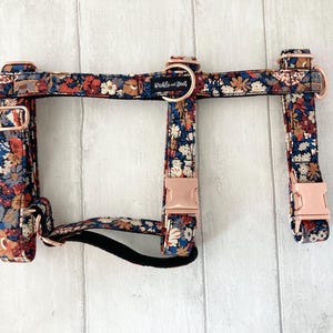 Puede incluir: Arnés para perro con estampado floral sobre fondo azul marino, con flores rojas, naranjas y blancas. Hebillas de metal oro rosa y correa negra. Se ve el nombre de la marca "Pickle and Dot".