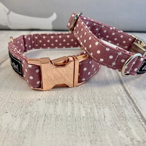 May include: Two dog collars. The collars are made of mauve fabric with white polka dots. One collar is buckled, and the other is unbuckled. The buckles are rose gold in colour. The collars have a black label with white text.