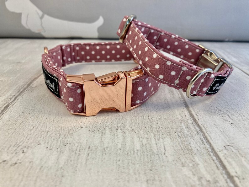 May include: Two dog collars. The collars are made of mauve fabric with white polka dots. One collar is buckled, and the other is unbuckled. The buckles are rose gold in colour. The collars have a black label with white text.
