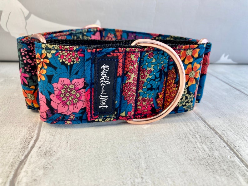 K&ouml;nnte beinhalten: Ein gemustertes Hundehalsband mit einem D-Ring und Beschl&auml;gen aus ros&eacute;goldfarbenem Metall. Das Halsband hat einen marineblauen Hintergrund mit rosa, blauen, orangefarbenen und gelben Blumen. Ein kleines marineblaues Etikett tr&auml;gt die Aufschrift "Pickle and Boot".