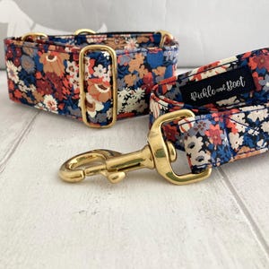 May include: A floral dog collar and matching leash set. The collar and leash feature a navy blue fabric with a pattern of red, orange, and white flowers. The hardware is gold-colored. The leash has a label that reads "Pickle and Boot".