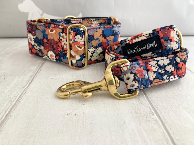 May include: A floral dog collar and matching leash set. The collar and leash feature a navy blue fabric with a pattern of red, orange, and white flowers. The hardware is gold-colored. The leash has a label that reads "Pickle and Boot".