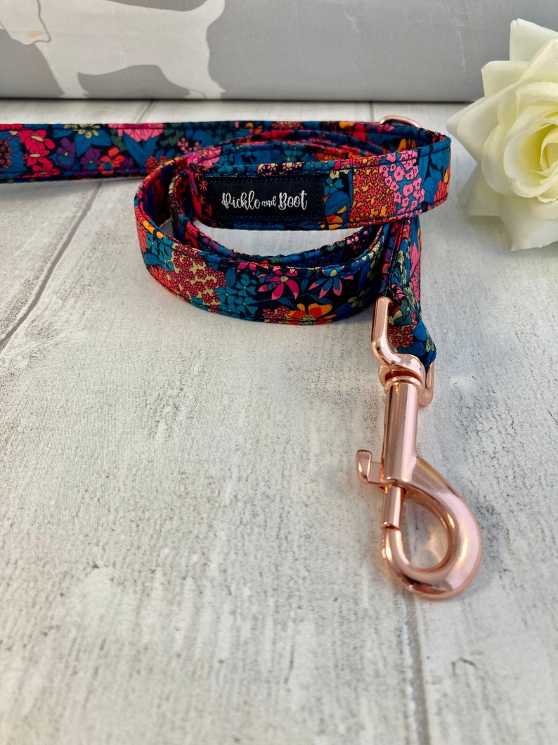 K&ouml;nnte beinhalten: Eine Hundeleine mit floralem Muster und einem ros&eacute;goldenen Karabinerhaken. Der Stoff hat einen marineblauen Hintergrund mit rosa, orangefarbenen und blauen Blumen. Das Etikett lautet "pickle and boot".