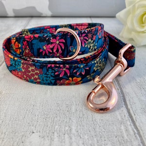 K&ouml;nnte beinhalten: Halsband und Leine f&uuml;r Hunde mit floralem Muster. Der Stoff hat einen marineblauen Hintergrund mit rosa, orangefarbenen und blauen Blumen. Die Beschl&auml;ge sind ros&eacute;goldfarben.