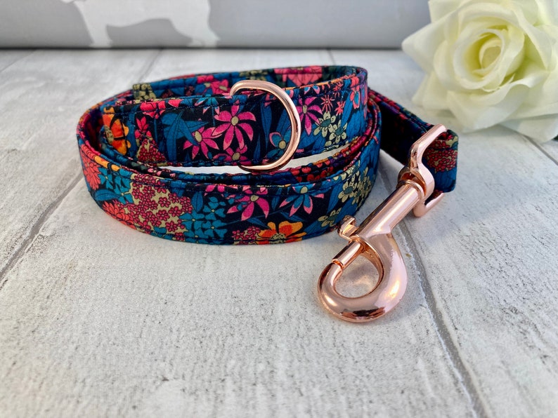 K&ouml;nnte beinhalten: Halsband und Leine f&uuml;r Hunde mit floralem Muster. Der Stoff hat einen marineblauen Hintergrund mit rosa, orangefarbenen und blauen Blumen. Die Beschl&auml;ge sind ros&eacute;goldfarben.