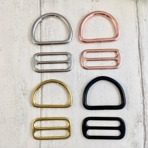 K&ouml;nnte beinhalten: Eine Auswahl an D-Ringen und Schiebeschnallen in Silber, Ros&eacute;gold, Gold und Schwarz. Die D-Ringe sind halbkreisf&ouml;rmig und die Schiebeschnallen haben zwei parallele Schlitze. Diese Metallbeschl&auml;ge sind f&uuml;r die Herstellung bestimmt.