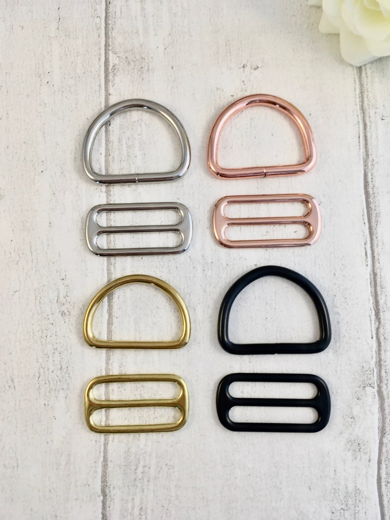K&ouml;nnte beinhalten: Eine Auswahl an D-Ringen und Schiebeschnallen in Silber, Ros&eacute;gold, Gold und Schwarz. Die D-Ringe sind halbkreisf&ouml;rmig und die Schiebeschnallen haben zwei parallele Schlitze. Diese Metallbeschl&auml;ge sind f&uuml;r die Herstellung bestimmt.