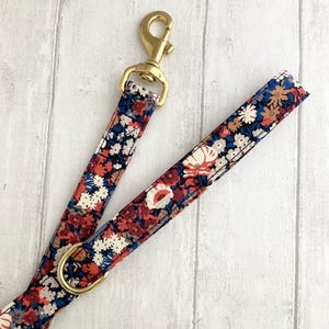 May include: A floral patterned dog leash with a brass clasp and ring. The leash features a repeating pattern of red, white, and blue flowers on a navy blue background. The brass hardware adds a touch of elegance to the design.