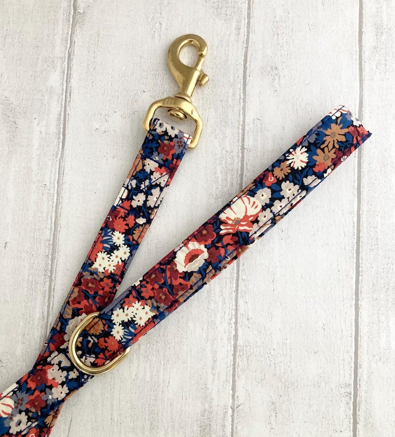 May include: A floral patterned dog leash with a brass clasp and ring. The leash features a repeating pattern of red, white, and blue flowers on a navy blue background. The brass hardware adds a touch of elegance to the design.