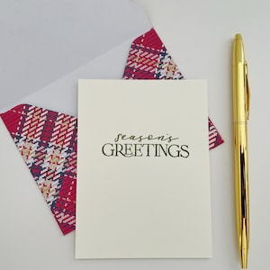 May include: A cream-colored greeting card with the text "season's GREETINGS" in green script, accompanied by a plaid envelope and a gold pen. The envelope's plaid pattern features red, white, and navy blue.