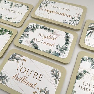 May include: A collection of rectangular cards with inspirational messages. Each card features a white background with gold text and botanical illustrations in green and gold. The cards have a light green border. Messages include "You're brilliant" and "So glad you exist."