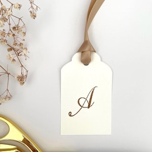 May include: A white gift tag with a gold ribbon and the letter "A" written in gold calligraphy.