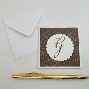 May include: A white envelope and a square card with a scalloped edge and the letter "G" in a script font. The card has a brown patterned background. A gold pen is below the card.