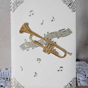 May include: A cream-colored card features a gold trumpet with musical notes. Decorative gold corner accents frame the card. The trumpet and musical notes are intricately cut out, creating a layered effect.