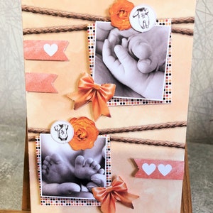 May include: A handmade baby-themed card with a peach background, featuring two black and white photos of a baby's hands and feet. The card is decorated with orange bows, paper flowers, and heart-shaped banners. Brown rope accents the design.