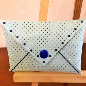 Peut inclure: Une pochette de couleur bleu clair en forme d'enveloppe avec un motif à pois et des accents bleus. La pochette a une fermeture à rabat et un bouton bleu.